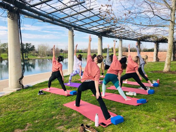 Group outdoor yoga class on colorful mats under a pergola beside a park pond, participants stretching in a warrior pose on a sunny day