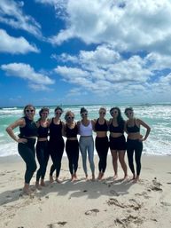 Eight women in athletic wear and heart-shaped sunglasses posing barefoot on a sunny sandy beach with turquoise waves and a bright blue sky dotted with fluffy clouds