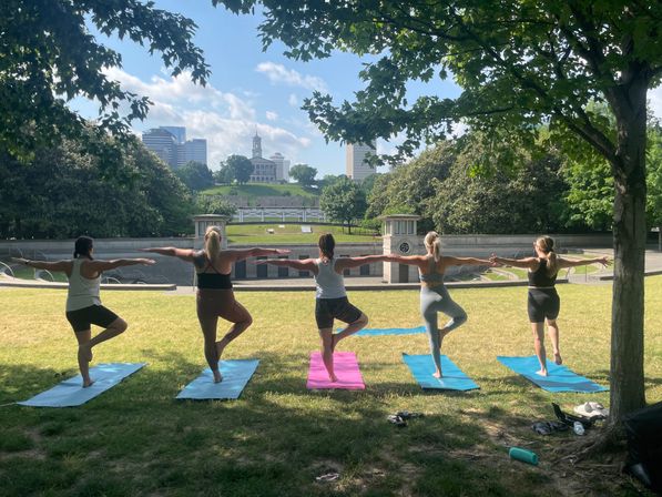 Group of five people doing tree-pose outdoor yoga on colorful mats in a shady city park, with a capitol-style building and downtown skyline on a sunny day.