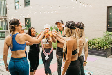 Women in workout clothes and caps toast with smoothies over yoga mats on a rooftop deck under string lights — outdoor urban fitness celebration.