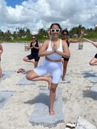 Group beach yoga on a sun-soaked tropical shore — woman in white activewear and sunglasses holding tree pose on a gray mat, friends practicing behind her with palm trees and blue sky in the background.