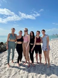 Group of six people in activewear standing barefoot on a sunny sandy beach by a turquoise ocean, smiling with a distant coastal skyline under a bright blue sky with scattered clouds.