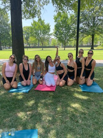 Eight women in activewear kneeling on colorful yoga mats under shade in a sunny city park, smiling and posing with a bottle of sparkling wine for an outdoor group fitness/gathering on a green lawn