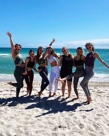 Seven friends in colorful activewear and heart-shaped sunglasses posing barefoot on a sunny turquoise-ocean beach, playfully jumping and striking peace signs on white sand under a clear blue sky.