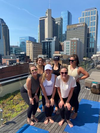 Six smiling women in athletic wear posing barefoot on yoga mats for a rooftop yoga session on a sunny day with a downtown skyline and clear blue sky behind them.