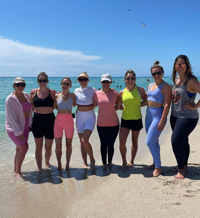 Eight women in bright athletic wear standing arm‑in‑arm on a sunny sandy beach shoreline with turquoise ocean and blue sky, kite and swimmers visible in the distance