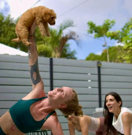 Woman in a teal sports bra holds a small curly-haired puppy aloft while in a side-plank during a sunny outdoor backyard yoga class, friends smiling nearby with palm trees and a fence in the background.