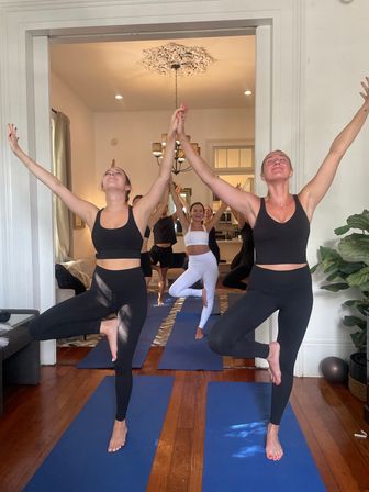 Sunlit home yoga class in a living-room studio: a group holding tree pose on blue mats, two women in black activewear clasping hands, hardwood floors, chandelier and houseplants.