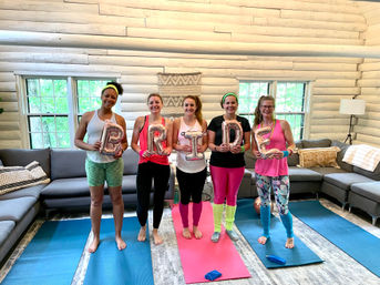 Five women in colorful workout gear pose on yoga mats inside a bright log‑cabin living room, holding rose‑gold letter balloons spelling BRIDE — playful indoor bachelorette yoga session.