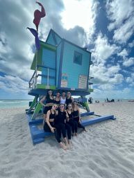 Eight women in black activewear wearing heart-shaped sunglasses pose on the sand in front of a bright turquoise-and-lime lifeguard tower with red and purple flags on a sunny, partly cloudy Miami beach