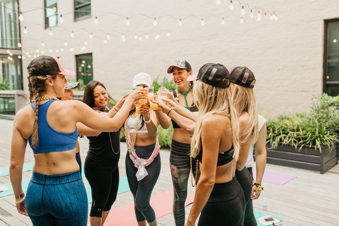 Women in athletic wear toasting with drinks on an urban rooftop deck with string lights and yoga mats — outdoor yoga/fitness celebration