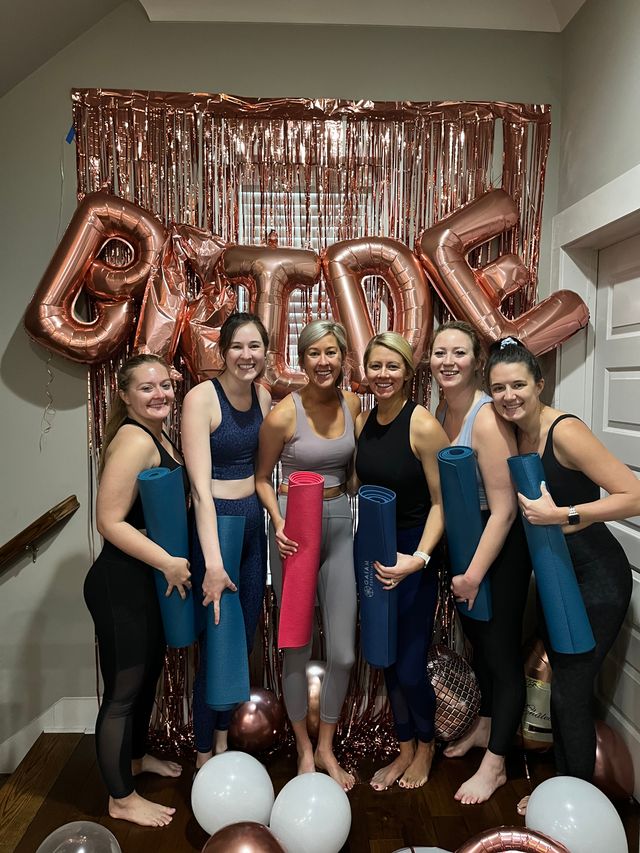 Six women in workout clothes holding yoga mats pose barefoot in an at-home bachelorette yoga yoga party with rose-gold metallic fringe backdrop, large rose-gold 'BRIDE' balloons, and scattered white and rose-gold balloons