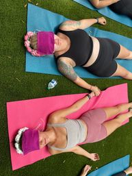 Two women holding hands while lying on pink and blue yoga mats on outdoor turf during a restorative yoga session, wearing purple eye masks and floral crowns