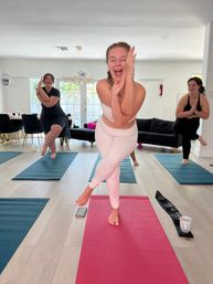 Three women smiling and balancing in eagle yoga pose on pink and blue mats in a bright sunlit living room — cheerful at-home group yoga session focused on fitness and wellness.