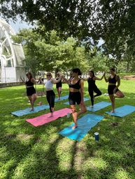 Group of women practicing tree pose on colorful yoga mats in a shaded city park near a white pavilion, bright green lawn and trees on a sunny day