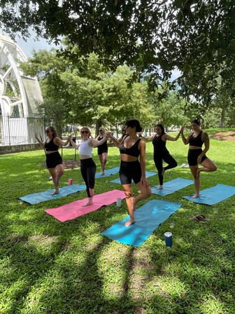 Group of women practicing tree pose on colorful yoga mats in a shaded city park near a white pavilion, bright green lawn and trees on a sunny day