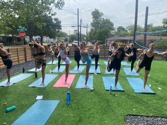 Energetic outdoor group yoga class on artificial turf in a suburban yard — participants balancing on one leg on colorful mats under an overcast sky.