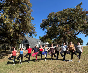 Outdoor yoga group of 11 participants holding tree pose hand-in-hand on a sunny grassy lawn in front of a country house beneath large oak trees