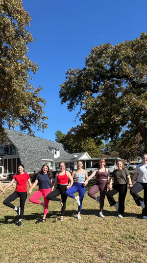 Outdoor yoga group of 11 participants holding tree pose hand-in-hand on a sunny grassy lawn in front of a country house beneath large oak trees