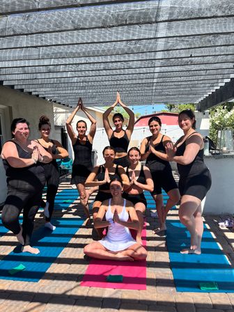 Sunlit outdoor yoga class on a backyard patio under a wooden pergola, women on colorful mats in prayer and tree poses with an instructor seated in white at the front.