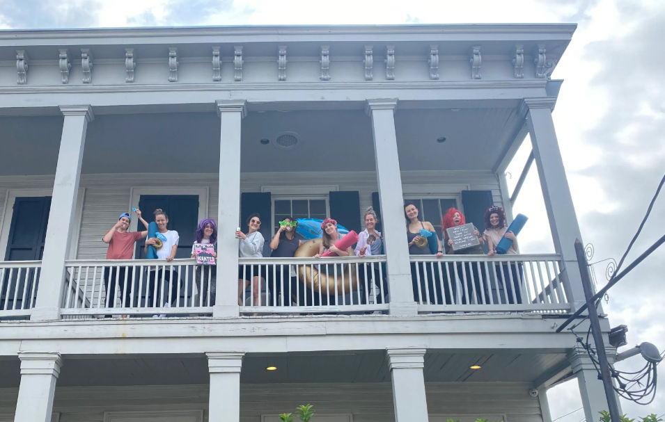 Group of friends laughing and posing on a second-floor white wooden balcony of a two-story house, holding colorful pool floats, yoga mats and party props against a cloudy sky.