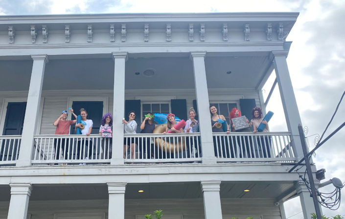 Group of friends laughing and posing on a second-floor white wooden balcony of a two-story house, holding colorful pool floats, yoga mats and party props against a cloudy sky.