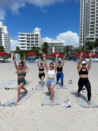 Group beach workout: five women in athletic wear doing lunges with arms raised on yoga mats on a sunny oceanfront beach, red umbrellas, palm trees and white high-rise condos under a bright blue sky.