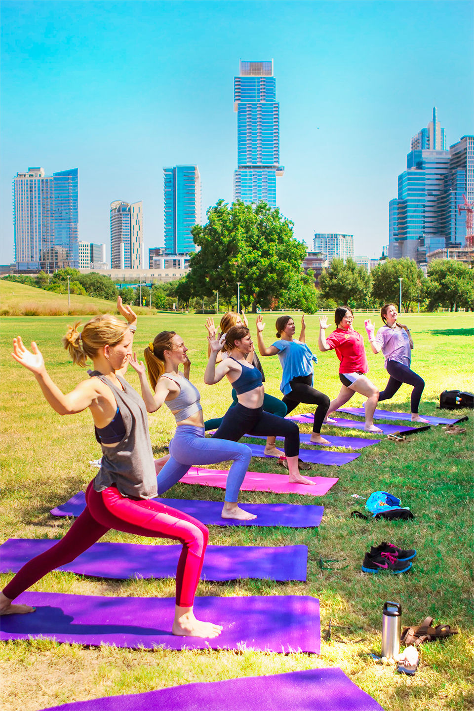 Sunny outdoor yoga class in an urban park — participants on purple mats practicing warrior poses with a downtown skyscraper skyline in the background.