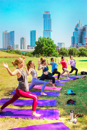 Sunny outdoor yoga class in an urban park — participants on purple mats practicing warrior poses with a downtown skyscraper skyline in the background.