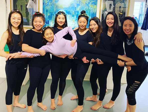 Group fitness/yoga studio photo of eight women in black activewear and floral headbands holding a smiling friend horizontally, barefoot on the studio floor with colorful abstract paintings behind them.