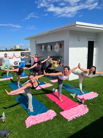 Rooftop yoga class on artificial turf under a bright blue sky — a group of women balancing on colorful mats with pink striped towels and flower balloon decorations on a sunny urban rooftop
