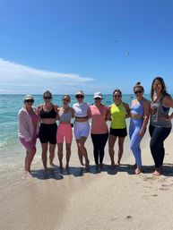 Group of nine friends in colorful activewear standing ankle-deep in turquoise ocean on a sunny beach with clear blue sky and a kite overhead