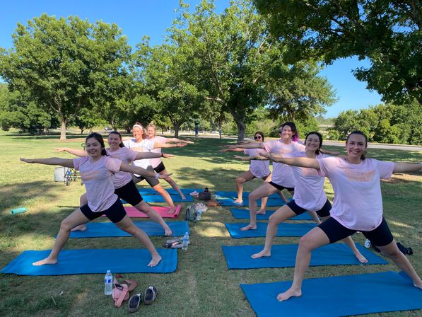 Group of women wearing matching pink shirts and black shorts smiling and holding Warrior II on blue mats on green grass during a sunny outdoor yoga class in a tree-lined park.