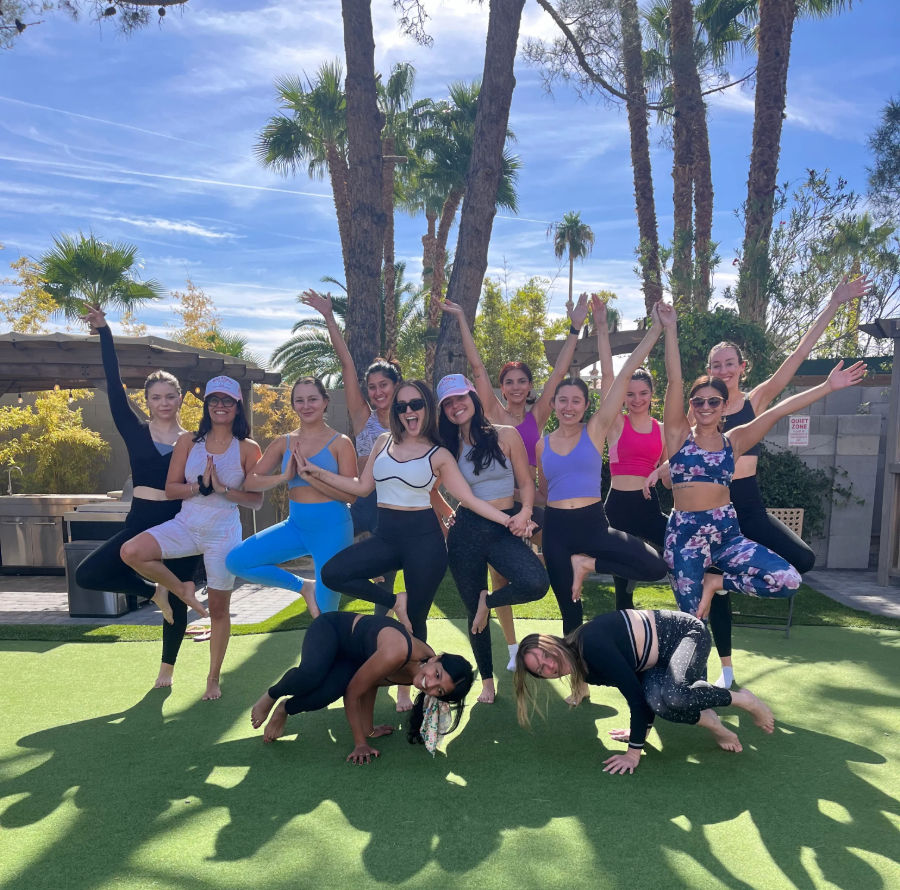 Large group of women in colorful activewear striking yoga and playful balancing poses on a sunny artificial lawn framed by palm trees and a bright blue sky.