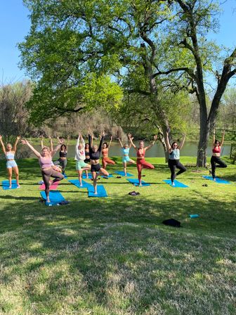 Group outdoor yoga class in a sunny riverside park: a dozen people on blue mats practicing tree pose on a grassy lawn beneath a large leafy tree beside a calm river.