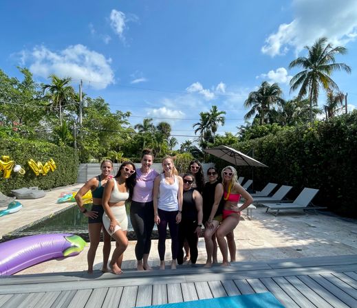Eight friends posing by a tropical backyard pool with palm trees, lounge chairs and inflatable floats under a bright blue South Florida sky — sunny pool party vibe.