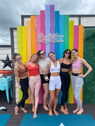 Six women in activewear posing barefoot on a rooftop yoga deck in front of a rainbow wooden wall with neon sign "Be A Voice Not An Echo" under a cloudy sky, one holding a bottle of sparkling wine.