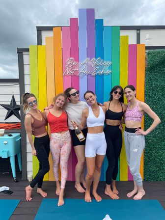Six women in activewear posing barefoot on a rooftop yoga deck in front of a rainbow wooden wall with neon sign "Be A Voice Not An Echo" under a cloudy sky, one holding a bottle of sparkling wine.