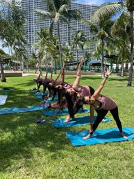 Group outdoor yoga class of women in matching tank tops doing triangle pose on blue mats in a sunny palm‑tree park with high‑rise buildings in the background and a child watching.