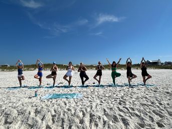Group beach yoga: people in tree pose on mats along a sunny white-sand shore under a clear blue sky with coastal dunes and houses behind them.