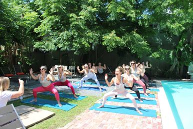 Energetic group of women doing an outdoor poolside yoga class on blue mats in a sunny backyard garden under leafy trees.