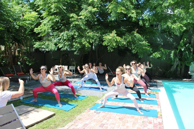 Energetic group of women doing an outdoor poolside yoga class on blue mats in a sunny backyard garden under leafy trees.