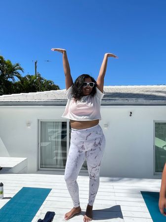 Person in sunglasses and white crop top with marble leggings stretching arms overhead on a sunny rooftop deck with yoga mats, palm trees and clear blue sky.