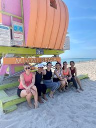 Six people in activewear sitting on sand beneath a colorful Miami Beach lifeguard tower (74th St), smiling on a sunny day with the ocean and clear blue sky in the background.