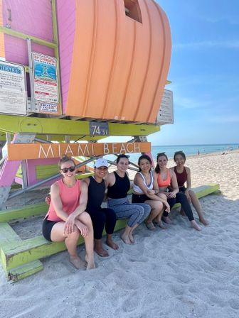 Six people in activewear sitting on sand beneath a colorful Miami Beach lifeguard tower (74th St), smiling on a sunny day with the ocean and clear blue sky in the background.