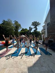 Outdoor poolside yoga class on blue mats, a group striking tree pose on a sunny suburban backyard patio beside a pool and brick house