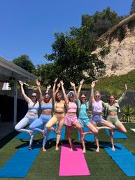 Seven women in pastel activewear practicing tree pose on yoga mats in a sunny backyard beside a rocky hillside and clear blue sky, outdoor group yoga session.