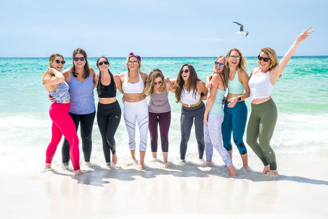 Nine women in colorful activewear laughing and posing at the water's edge on a sunny turquoise beach with a seagull flying overhead.