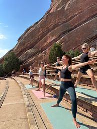 Sunny outdoor yoga class on colorful mats at a red sandstone amphitheater in Colorado, participants striking Warrior II poses against towering rock walls and blue sky