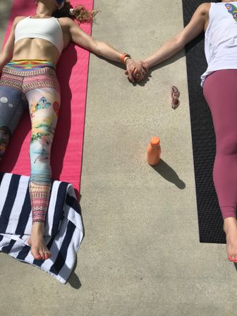 Sun-soaked outdoor yoga on a concrete patio: two people lying on pink and black mats holding hands, colorful leggings, sunglasses and orange water bottle nearby.